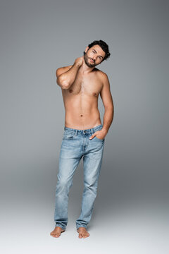 full length of good-looking and muscular man in blue jeans posing with hand in pocket on grey