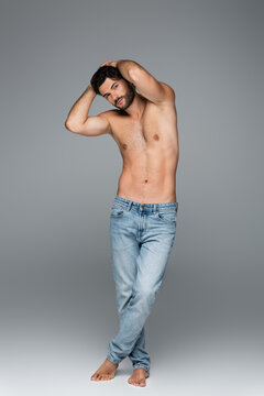 full length of good-looking and muscular man in jeans looking at camera while posing on grey