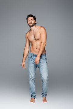 full length of good-looking and muscular man in jeans looking at camera on grey