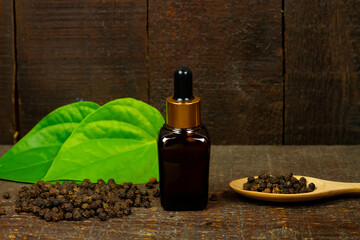 Black pepper essential oil in glass bottle with peppercorn on wooden spoon and green leaf on wooden background.