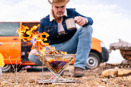 Mature man crouching near wood burning stove while camping on ground