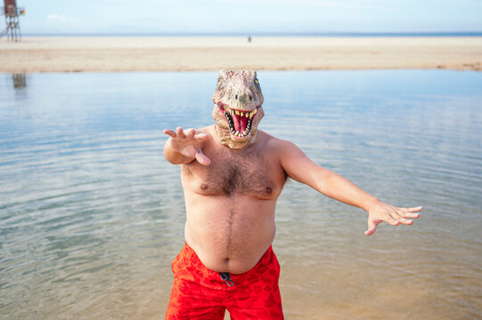 Shirtless man wearing lizard mask gesturing at beach