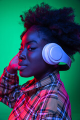 Close-up young girl, student listening to music in headphones isolated on dark green studio background in purple neon light.