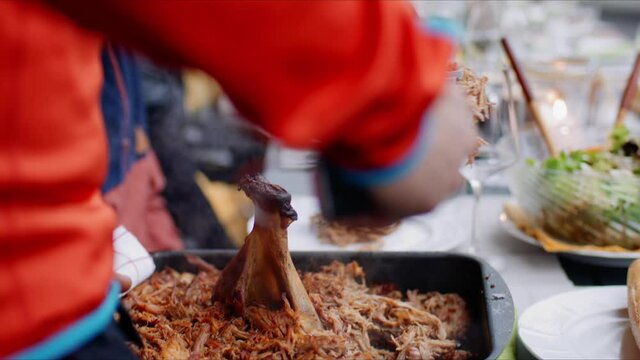 Putting pulled pork out of pan into bread roll on plate with many plates and salad in background
