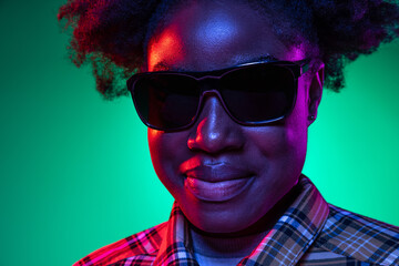 Close-up face of dark skinned young girl in sunglasses isolated on dark green studio background in purple neon light.