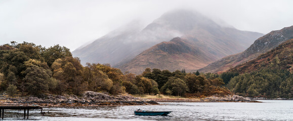 Small boat on a tranquil loch with the Glencoe mountain range in the distance - obrazy, fototapety, plakaty