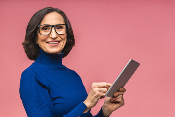 Portrait of smiling beautiful senior aged mature woman using tablet computer, isolated over pink background. Copy space for text.