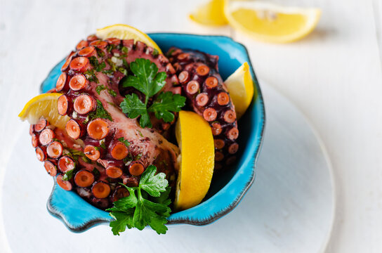 Healthy Mediterranean delicacy. Boiled Octopus with lemon and parsley in a blue bowl on white background. Selective focus, copy space