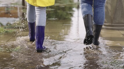 girls in raincoats and rubber boots walk along road flooded with torrential rains, their feet walk...