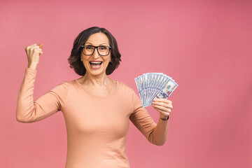 Happy winner! Image of a mature senior happy old woman standing isolated over pink background, holding money. - 473815943