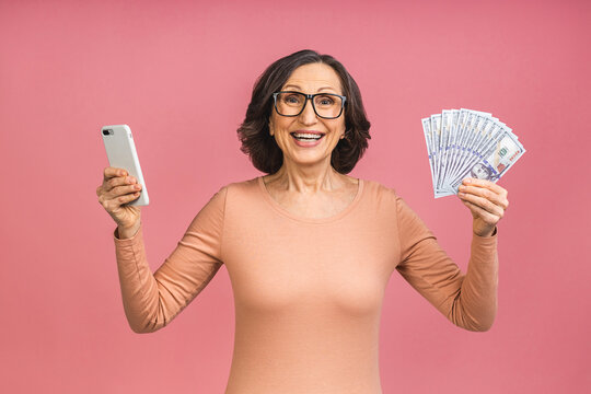 Happy winner! Image of mature aged happy senior woman standing isolated over pink background wall looking at camera holding money using mobile phone.