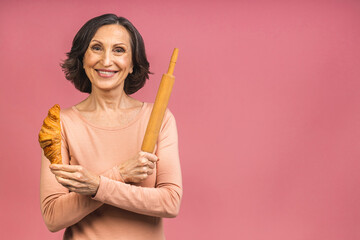 Senior mature aged woman baker holding a rolling pin and croissant, isolated on pink background.