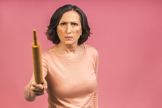 A very angry senior mature aged lady holding a rolling pin and threatening to whack someone with it (her husband?). Isolated on pink background.