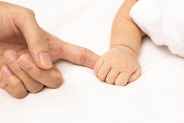 Little baby boy in grey pajamas holding mother's hand on the bed, close up