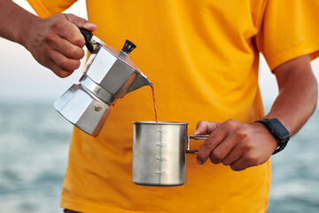Man in an orange t-shirt pours coffee from vintage geyser coffee maker into a steel mug against the...