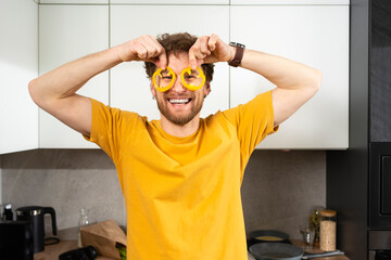 Smiling man holding bell pepper slices on eyes in kitchen at home