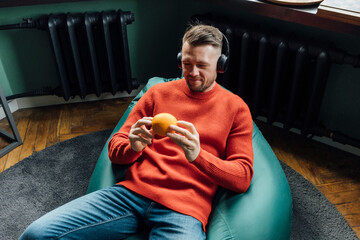 Smiling male entrepreneur with headphones looking at orange fruit while sitting on bean bag