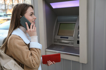 Young woman talking by mobile phone while using cash machine for money withdrawal outdoors