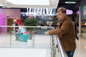 An adult blond man in corduroy jacket is bored at the mall waiting for his wife.