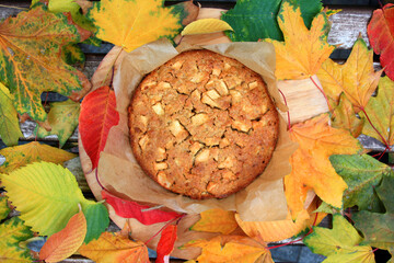 Among the large number of yellow, orange, red, green, colorful leaves on a wooden stand lies a small tasty round cake. Delicious apple pie covered with cracks. Autumn atmosphere.