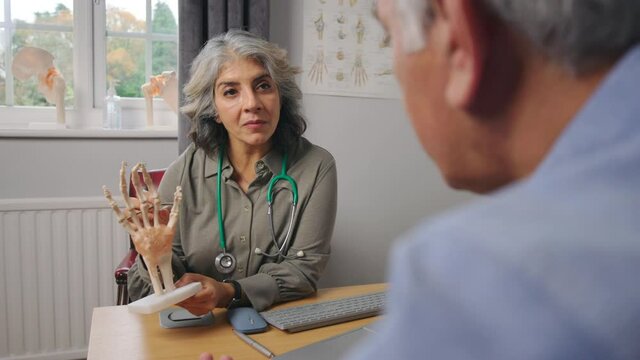 Mature Female Doctor Meeting With Male Patient Discussing Joint Pain In Hand Using Anatomical Model