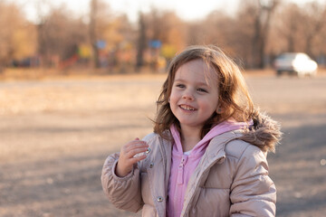 Fototapeta premium Beautiful red-haired little girl in a jacket with a fluffy hood smiles on the background of an autumn street