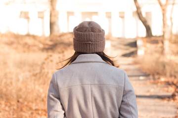 A girl with long dark hair in a coat and a knitted hat stands with her back to the camera against...