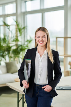 Smiling Blond Female Intern Holding File While Standing With Hand In Pocket At Office