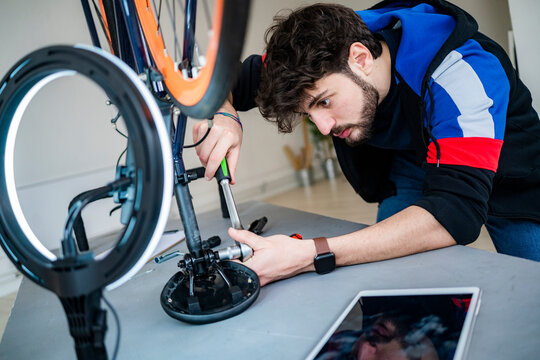 Concentrated young man fastening bicycle seat with wrench while filming at home