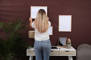 Woman hanging empty frame on brown wall indoors, back view. Mockup for design