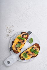 Scrambled eggs with fried mushrooms and basil on bread on white table background. Homemade breakfast or brunch meal. Scrambled eggs and mushrooms sandwiches. Top view with copy space