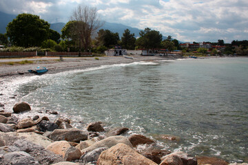 Rocky seashore. In the distance, the city and mountains are visible. Sun glare on the water. Overcast weather. Greece, Europe.
