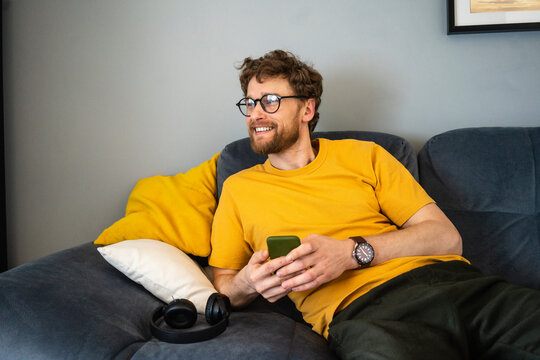 Smiling mid adult man with eyeglasses and mobile phone looking away while sitting on sofa