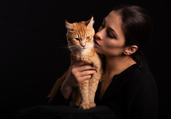 Beautiful make-up woman hugging and kissing with love her red maine coon kitten. Closeup portrait. Art on black background