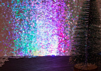 Colored background with free space for text or logo. New Year's atmosphere. Selective focus and blurry multi-colored bokeh. Small decorative Christmas tree in the foreground.