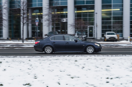 car BMW 5 Series E60 n the winter city street. Side view of black sedan vehicle in motion against blurred urban background