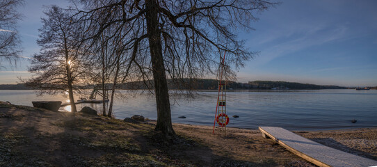 Pale Winter solstice reflex in the calm lake Mälaren, beach with jump tower and life saver at an...
