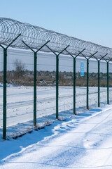 Iron fence with barbed wire in a snow-covered winter field against a blue sky. Fenced guarded territory, no access. Area protection and counteraction to illegal entry concept