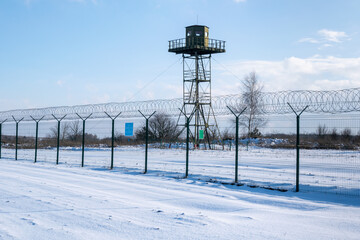 Border fence with barbed wire in front of the surveillance tower in a snow-covered field against a...