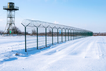 Border fence with barbed wire in front of the surveillance tower in a snow-covered field against a...