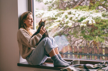 Young cheerful girl sitting at home on the windowsill in a warm sweater playing with a gray cat on...