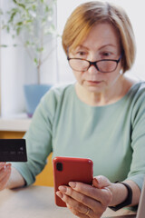 Close up smartphone in hand, worried confused senior woman online buyer holding credit card,looking at mobile, sitting indoor. Debt problems, insecure online payment, failed transaction, losing money