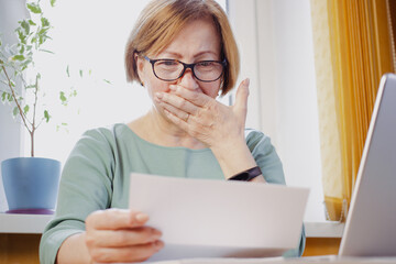 Shocked worried mature woman reading paper document or postal letter feeling frustrated by unexpected news sitting at home indoor. Received bank account balance or bad notice, fine, bankruptcy