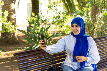 Young Muslim woman wearing hijab sitting on park bench taking a selfie