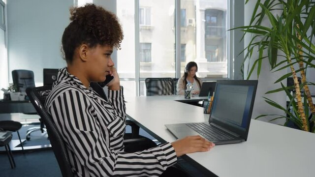African American girl wearing striped shirt using laptop and talking on phone in open space office, Asian coworker with headphones on background. Arc shot multiethnic employees at work