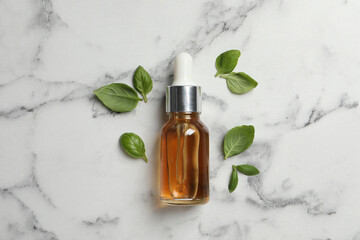 Bottle of essential basil oil and fresh leaves on white marble table, flat lay