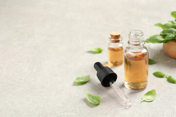 Bottles of essential basil oil and fresh leaves on light grey table, space for text