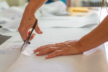 Hands of a woman dressmaker cutting a white fabric with professional scissors. Clothing and textile design concept.