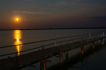 Fototapeta premium A wooden pedestrian bridge with railings runs along the lake, around the shallow water. There are bright stars and the moon in the sky.