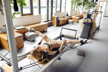 Sporty young woman exercising on pilates reformer machine bed in modern gym studio